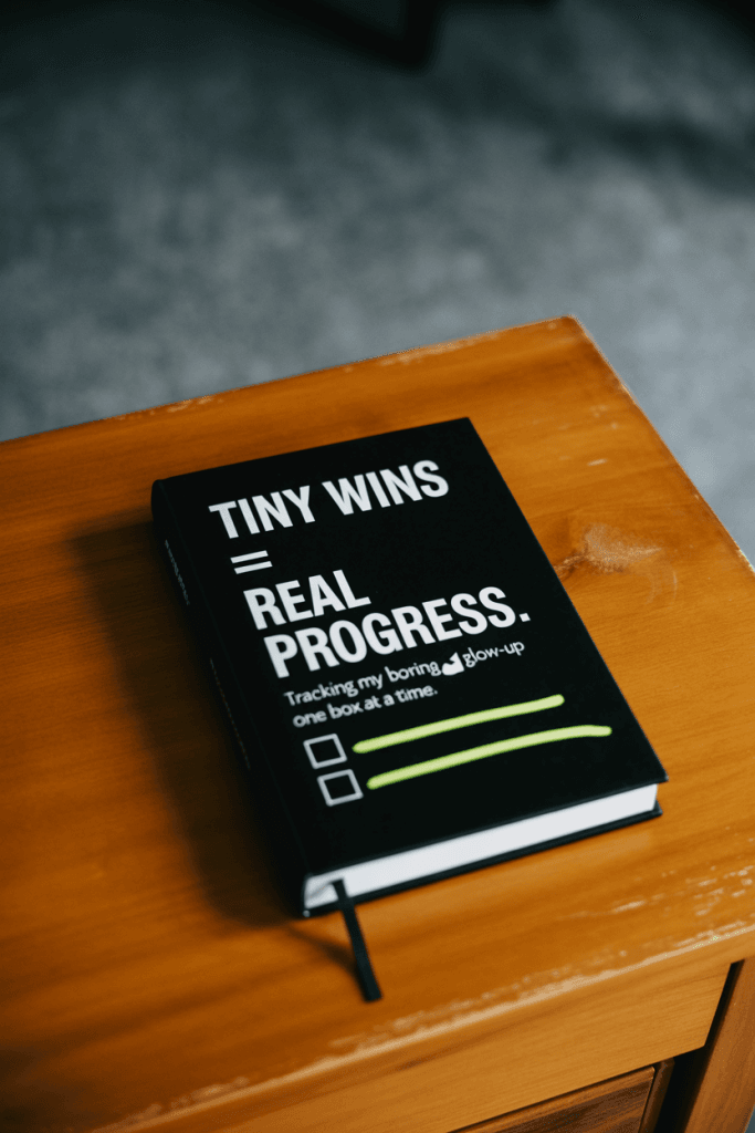 Minimalist journal layout on a wooden desk with soft shadows. A white text overlay says “Tiny Wins = Real Progress.” A checklist and highlighter rest on the side for a simple, cozy productivity mood.