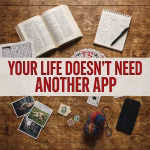 Colourful flat lay of a paperback, notebook, crossword, playing cards and printed photographs with the words “Your Life Doesn’t Need Another App”.